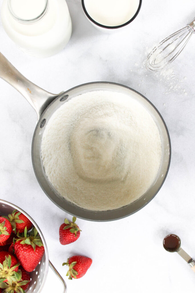 Flour and cornstarch mixed together in a gray sauce pan on a marble countertop surrounded by strawberries, milk and vanilla extract. 