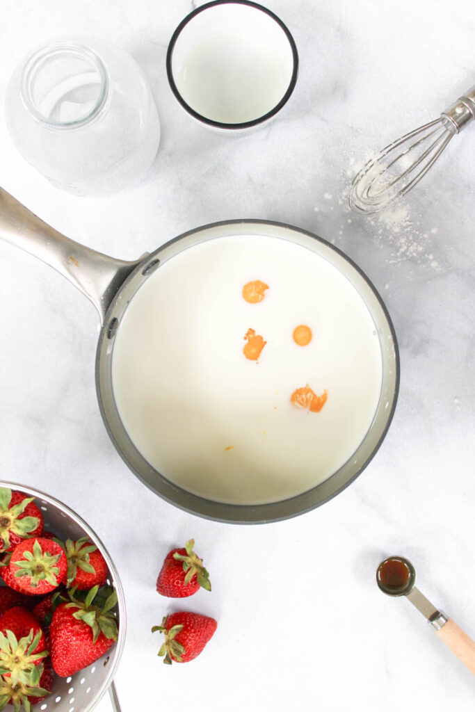 egg yolks, milk, and half and half are mixed together with sugar and cornstarch in a gray sauce pan on a marble countertop surrounded by strawberries, vanilla extract and a whisk. 