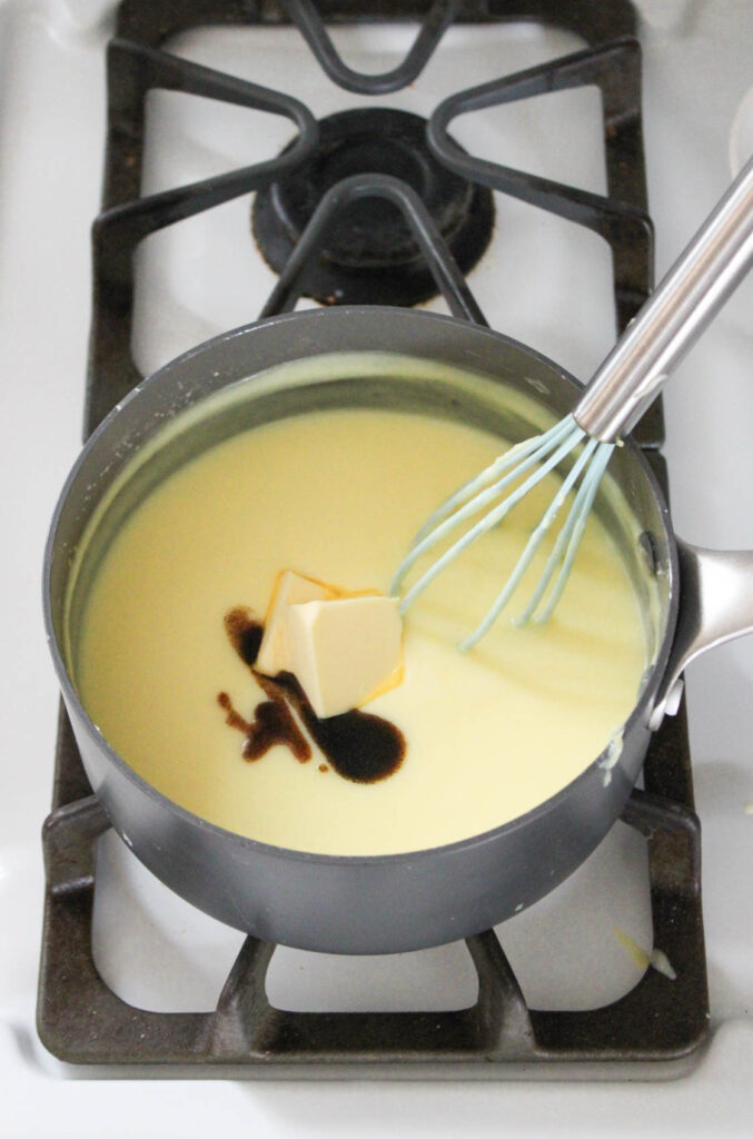 butter and vanilla extract are added to custard being cooked in a gray pan with a whisk on the stove. 