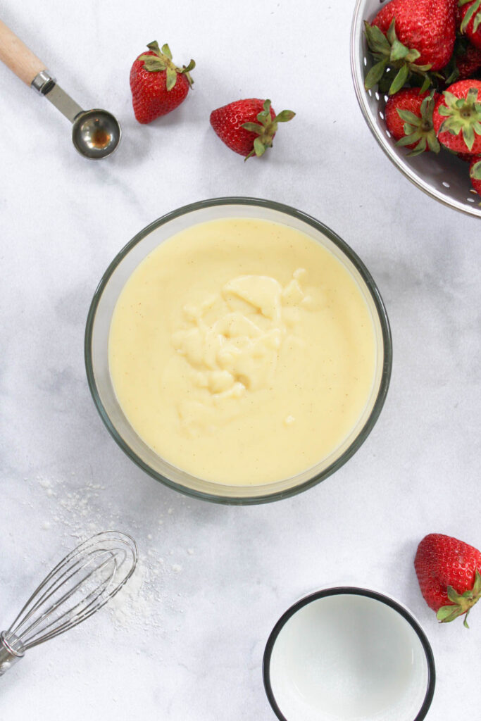 vanilla custard is in a glass bowl on a marble countertop surrounded by strawberries. 