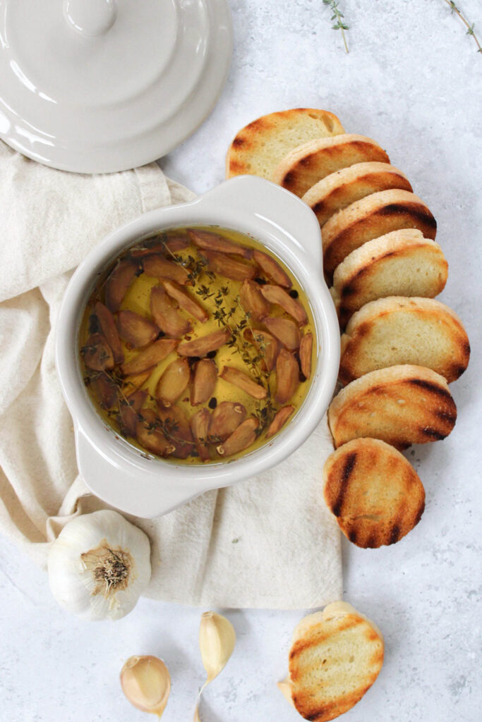 garlic confit in a small tan bowl with handles surrounded by grilled bread on a stone countertop. 