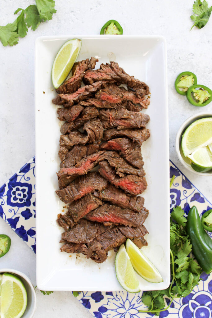 Ranchera meat with a carne asada marinade on a long plate surrounded by sliced limes, jalapeños, and cilantro.