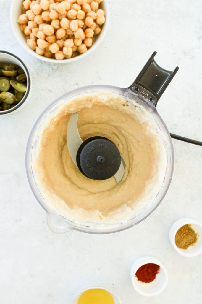 Tahini and lemon juice blended in a food processor with garbanzo beans, jalapeños, and spices on the counter next to it.  