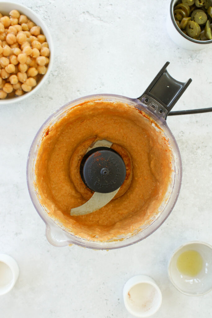 Tahini, lemon juice, garlic, spices, and olive oil blended in a food processor with chickpeas and jalapeños on the counter next to it. 