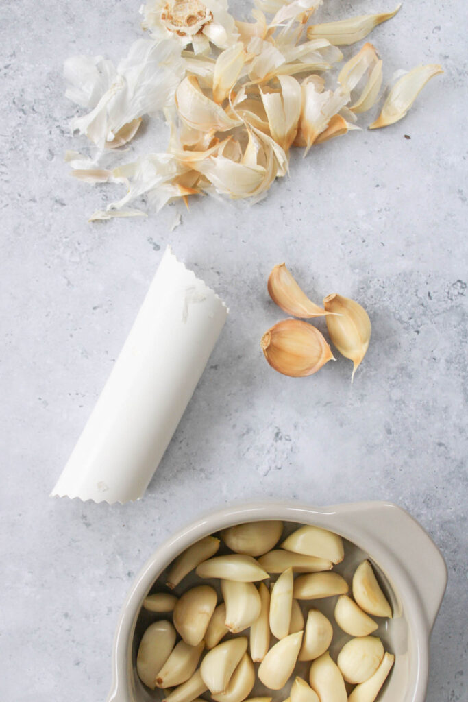 peeled garlic in a small tan bowl with handles and garlic skins on a stone counter top. 