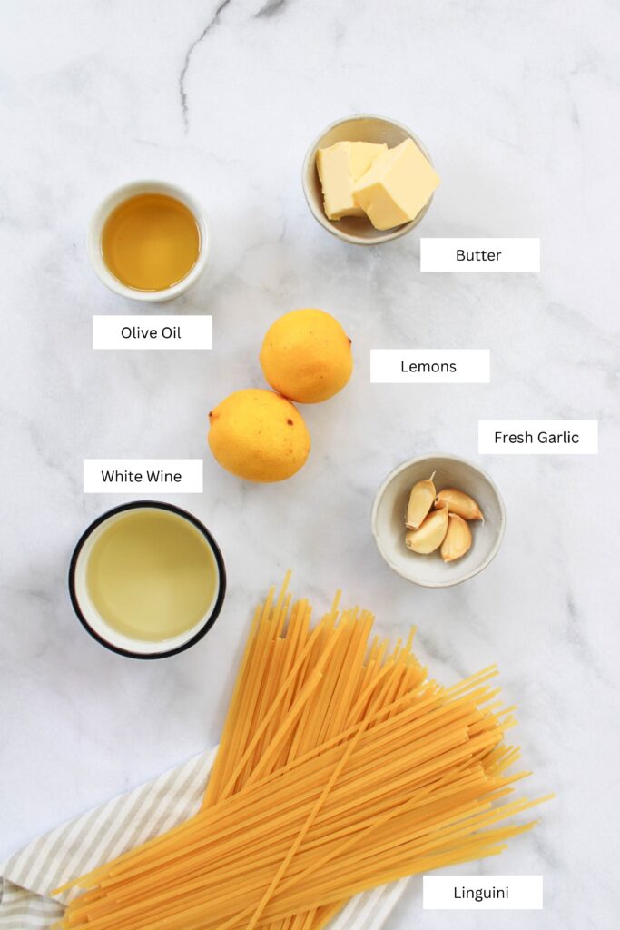 Garlic lemon white wine pasta ingredients including lemons, garlic, white wine, butter, olive oil, and pasta on a marble counter top. 