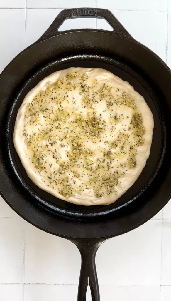 pizza dough topped with garlic olive oil and herb sauce in a cast iron skillet 
