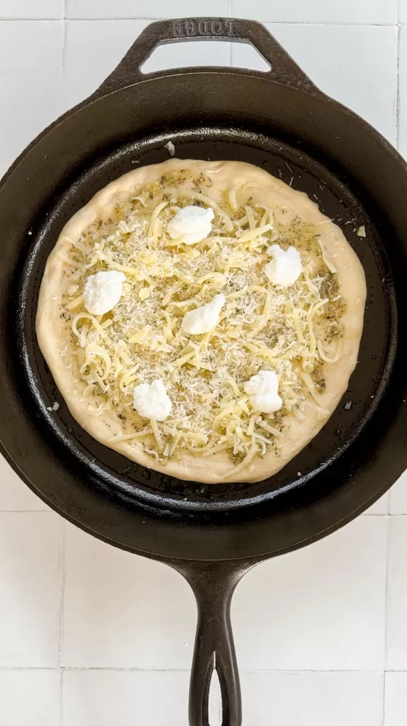 Pizza dough topped with garlic olive oil sauce, mozzarella cheese, parmesan cheese, and ricotta cheese in a cast iron skillet. 
