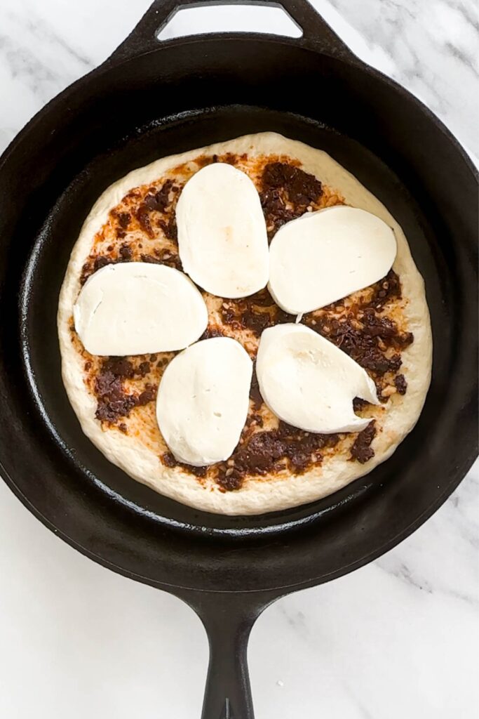 Pizza dough topped with sun-dried tomato spread and sliced fresh mozzarella in a cast iron skillet. 