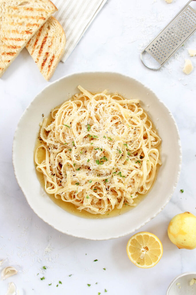 Garlic lemon white wine pasta sauce on top of pasta surrounded by grilled bread, lemons, and parmesan cheese. 